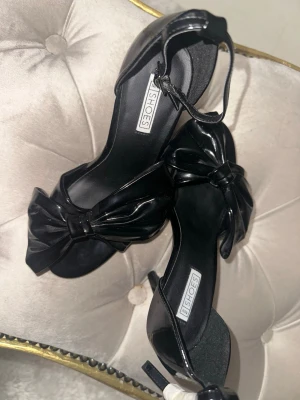 Svarta lack klackar med rosett - Säljer ett par svarta klackar i lackmaterial från NELLY SHOES. Skorna har öppen tå, hög smal klack och en stor dekorativ rosett framtill. Rem runt ankeln med spänne för extra stöd. Perfekta för dig som vill ha en elegant och trendig look.