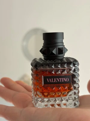 Valentino Eau de Parfum Intense - Säljer en lyxig parfym från Valentino i en cool, fyrkantig glasflaska med nitar och svart kork. Flaskan har en mörk orange vätska och en etikett med Valentino-loggan. Perfekt för dig som gillar exklusiva dofter och snygg design.