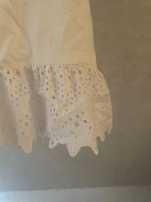 Vit topp med spetsvolang - Söt vit topp med korta ärmar och rund hals. Nederdelen har en bred volangkant i broderad spets med hålmönster och vågig avslutning. Perfekt för dig som gillar romantiska detaljer och en feminin stil. Använd 1 gång 