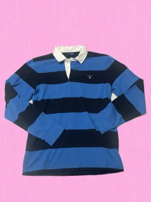 Randig rugbytröja från Gant stl M - Klassisk långärmad rugbytröja från Gant med breda blå och marinblå ränder samt vit krage. Tröjan har knappslå vid halsen och broderad logga på bröstet. Tillverkad i mjuk bomull som känns skön mot huden. Perfekt för en sportig och avslappnad look. Den är egentligen helt ny testats på ca 2-3 gånger men aldrig använts dock lite smink på kragen när jag testade syns nästan inte alls (se de 3 sista bilderna). Nypris runt 1500kr