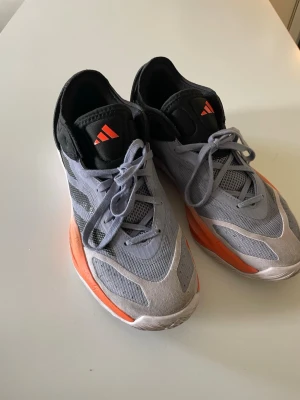 Adidas Adizero grå och orange sneakers - Snygga Adidas Adizero sneakers i grått med svarta och orange detaljer. Skorna har meshpaneler för bra ventilation, klassisk snörning och låg profil. Perfekta för dig som gillar sportig stil och vill sticka ut med färg. Adidas-logga på plösen och sidan.