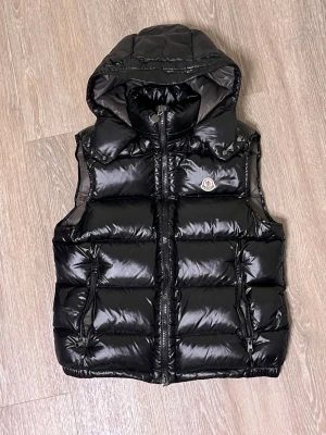 Svart dunväst från Moncler med huva - Snygg svart dunväst från Moncler med glansig finish, avtagbar huva och två dragkedjefickor framtill. Västen har klassisk Moncler-logga på bröstet och quiltad design. Perfekt för lager-på-lager och streetstyle.