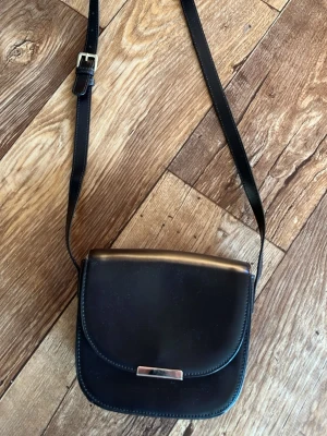 Mörkbrun  Wera axelväska i läderlook - Stilren svart axelväska i slät läderimitation med justerbart axelband och silverfärgat spänne framtill. Väskan har en klassisk, rundad form och enkel design som passar till många outfits.