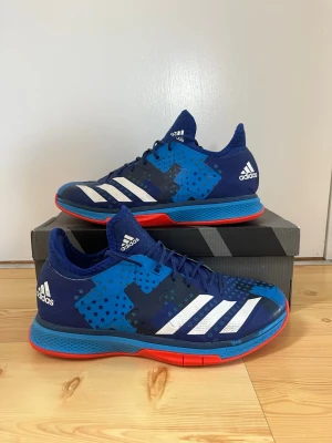 Blå Adidas inomhusskor - Säljer ett par snygga Adidas inomhusskor i blått med vita ränder och detaljer i svart. Skorna har en röd och blå sula som ger grymt grepp på planen. Perfekta för handboll eller innebandy. Ovandelen är i mesh och syntet för bra ventilation och komfort.