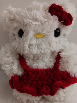 Virkad Hello Kitty - Söt vit plyschkatt med stora svarta ögon, gul nos och en röd klänning samt röd rosett på huvudet. Perfekt som dekoration eller present till någon som gillar mjuka gosedjur och kawaii-stil.