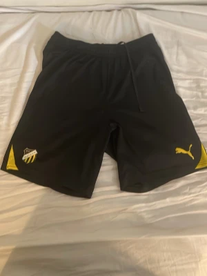 Svarta fotbollsshorts från Puma - Snygga svarta shorts från Puma med gul logga och klubbmärke på benen. Tillverkade i lätt och skönt polyestermaterial, perfekta för fotboll eller träning. Elastisk midja med snörning för bästa passform och gula detaljer på sidorna. 