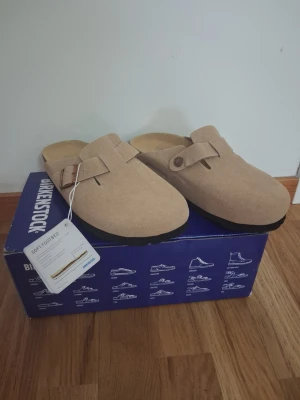 Beige Birkenstock tofflor i mocka - Säljer ett par snygga beige tofflor från Birkenstock i mjuk mocka med justerbar rem och metallknapp. De har en klassisk slip-in design och mjuk fotbädd för extra komfort. Perfekta för dig som gillar stilrena och bekväma skor.