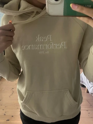 Beige hoodie från Peak Performance - Snygg beige hoodie från Peak Performance med broderad logga på bröstet. Klassisk känguruficka framtill och justerbar huva med dragsko. Mjuk bomullsblandning som är skön att ha på sig. Perfekt för en avslappnad och stilren look.