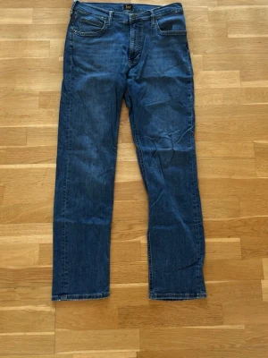 Blå raka jeans från Lee - Säljer ett par klassiska blå jeans från Lee med rak passform och fem fickor. Jeansen har normal midja och är tillverkade i slitstark denim. Perfekta för dig som gillar en tidlös och enkel stil.