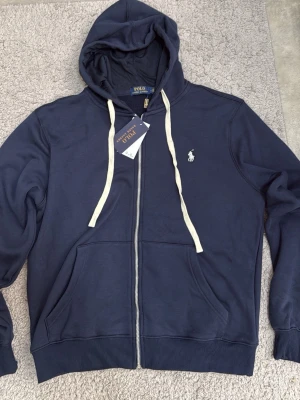 Marinblå hoodie från Polo Ralph Lauren - Snygg marinblå hoodie från Polo Ralph Lauren med dragkedja, vit logga broderad på bröstet och vita snören i huvan. Tillverkad i mjuk bomullsmix och har två fickor framtill. Perfekt för en avslappnad och stilren look.