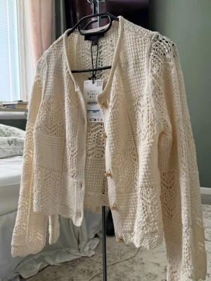 Krämvit virkad kofta från Zara - Trendig krämvit kofta från Zara i virkad design med snyggt hålmönster. Koftan har rund halsringning, långa ärmar och knappar framtill. Perfekt att slänga över en topp för en chill och stilren look. Materialet känns lätt och luftigt.
