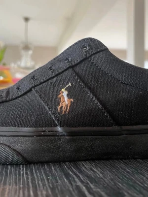 Svarta Polo Ralph Lauren sneakers - Snygga svarta sneakers från Polo Ralph Lauren med klassisk broderad logga på sidan. Skorna har en slitstark canvas-ovandel och en brun gummisula med POLO-tryck undertill. Perfekta för en clean och stilren look.