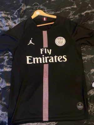 Psg tröja - Säljer en svart PSG matchtröja i samarbete med Jordan och Fly Emirates. Tröjan har korta ärmar, rund hals och en vertikal rand i ljusgrått framtill. PSG-logga och Jordan-logga på bröstet, samt Fly Emirates-tryck. Tillverkad i lätt och snabbtorkande material.