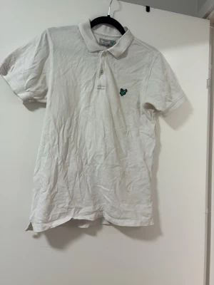 Vit pikétröja med broderad logga - Klassisk vit pikétröja från Lyle & Scott med korta ärmar och krage. Tröjan har knappar framtill och en grön broderad logga på bröstet. Tillverkad i bomull för en bekväm känsla och stilren look.