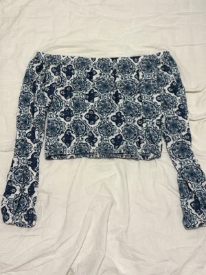 Offshoulder topp med blått mönster - Trendig offshoulder topp med lång ärm och blått, nästan bohemiskt mönster på vit botten. Croppad modell med resår upptill och nedtill för en snygg passform. Perfekt för dig som gillar unika detaljer och vill sticka ut.