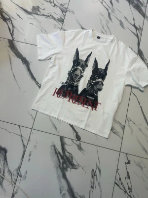 Vit Represent t-shirt med hundmotiv - Säljer en vit t-shirt från Represent med tryck av två svarta hundar på framsidan och texten 'REPRESENT' i rött. T-shirten har normal passform och korta ärmar. Perfekt för dig som gillar streetwear och vill sticka ut med ett coolt motiv. Pris kan diskuteras