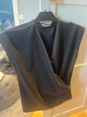 Svart omlottopp från Besilo - Snygg svart omlottopp från Besilo med djup v-ringning och ärmlös design. Toppen har en elegant drapering framtill och passar perfekt till många olika stilar. Tillverkad i ett mjukt och följsamt material.