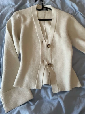 Vit cardigan från Gina tricot  - Snygg krämvit kofta med v-ringning och två stora, marmorerade knappar framtill. Koftan har långa ärmar med breda muddar och en lätt figursydd passform. Perfekt att slänga över en topp för en clean look. Använd ca 2 gånger säljer pga att den inte kommer till användning.