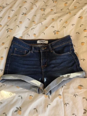 Mörkblå jeansshorts från ONLY - Snygga mörkblå jeansshorts från ONLY med uppvikta ben. Shortsen har normal passform och är perfekta för sommardagar. Gjorda i mjuk bomullsdenim med kontrastsömmar och knappgylf.