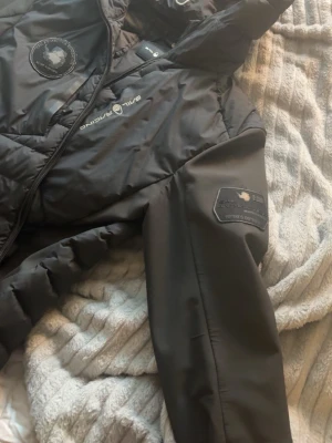 Svart pufferjacka Sail Racing - Säljer en svart pufferjacka från Sail Racing med patch på ärmen och logga på bröstet. Jackan har quiltad design, hög krage och dragkedja framtill. Materialet är glansigt och jackan har en sportig look med detaljerade sömmar och emblem.