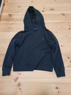Svart hoodie från Polo Ralph Lauren - Svart hoodie från Polo Ralph Lauren med klassisk huva och känguruficka. Liten röd broderad logga på bröstet. Mjuk bomullskänsla och ribbade muddar vid ärmslut och nederkant. Perfekt för en chill och stilren look.