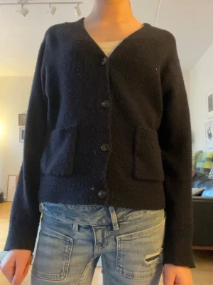 Mörkblå kofta med fickor och knappar - Mysig mörkblå kofta med v-ringning och fyra knappar framtill. Två stora fickor på framsidan ger en avslappnad look. Perfekt att slänga över en t-shirt eller linne för en enkel och stilren outfit.