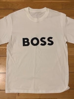 Vit BOSS t-shirt med tryck - Snygg vit t-shirt från BOSS med stort svart tryck på bröstet. Klassisk rund halsringning och korta ärmar. Tillverkad i mjuk bomull för en skön känsla och enkel stil.