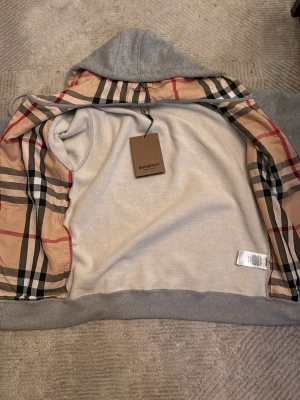 Grå hoodie från Burberry med dragkedja - Snygg grå hoodie från Burberry med klassiskt rutigt foder på insidan och huva. Dragkedja framtill, fickor och ribbade muddar. Perfekt för dig som vill ha en stilren och exklusiv look. Materialet är mjukt och bekvämt, och huvan har justerbara snören.