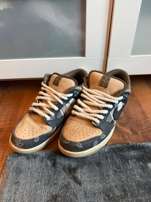 Nike SB Dunk Low Travis Scott - Nike SB Dunk Low Travis Scott med beige mocka och mörkblå paisleymönstrade paneler. Skorna har vita, tjocka rep-liknande skosnören och en klassisk låg siluett. Swoosh-logga på sidan och perforerad tåbox för extra ventilation.