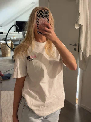 Vit Barbie The Icon t-shirt - Säljer en vit t-shirt från Barbie med trycket 'Barbie The Icon Since 1959' och en stor färgglad Barbie-hus illustration på ryggen. T-shirten har korta ärmar och en klassisk passform. Perfekt för dig som älskar retro och popkultur!