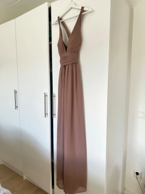 Lång brun omlottklänning med v-ringning - Säljer en elegant långklänning i brun nyans med omlottdesign och djup v-ringning. Klänningen har breda axelband och markerad midja med draperade detaljer. Perfekt för dig som vill ha en stilren och feminin look. Prislapp sitter kvar. 