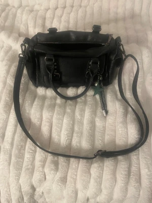 Svart handväska med axelrem - Snygg svart handväska i skinnimitation med både handtag och justerbar axelrem. Väskan har silverfärgade detaljer, dragkedja upptill och en dekorativ nyckelring med stjärna. Perfekt storlek för det viktigaste och flera fack inuti.