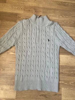 Ljusgrå kabelstickad tröja Polo Ralph Lauren - Säljer en ljusgrå kabelstickad tröja från Polo Ralph Lauren med halv dragkedja och hög krage. Tröjan har det klassiska broderade logotypen på bröstet och ribbade muddar vid ärmslut och nederkant. Perfekt för lager på lager och riktigt snygg till jeans.