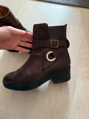 Bruna läderstövletter med spänne - Snygga bruna stövletter i läder med dekorativt spänne och silverfärgad ringdetalj på sidan.