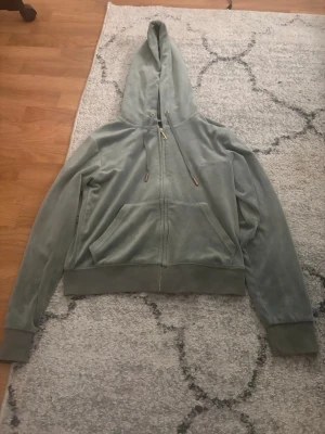  juicy couture tröja - Mjuk grön juicy couture tröja med dragkedja framtill och stora fickor. Modellen har huva med dragsko och ribbade muddar vid ärmslut och nederkant. Perfekt för en avslappnad och trendig look.