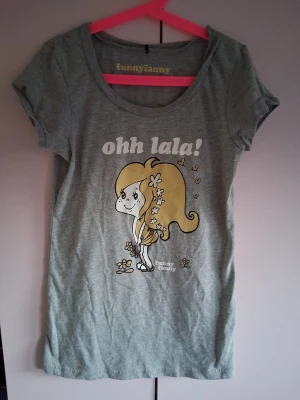Grå Funny Fanny t-shirt med tryck - Snygg grå t-shirt från Funny Fanny med ett gulligt tryck av en tecknad tjej och texten 'ohh lala!' framtill. T-shirten har rund halsringning och korta ärmar. Perfekt för dig som gillar roliga och unika motiv.