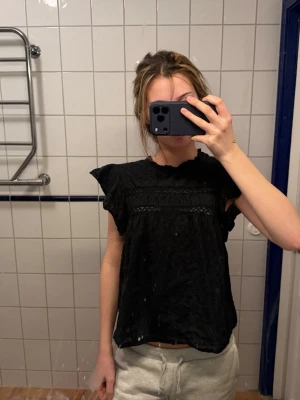 Svart blus från Vero Moda - Säljer en svart blus från Vero Moda med korta volangärmar och diskret prickigt mönster i tyget. Blusen har en lös passform och fina spetsdetaljer upptill. Perfekt att styla med både jeans och kjol.