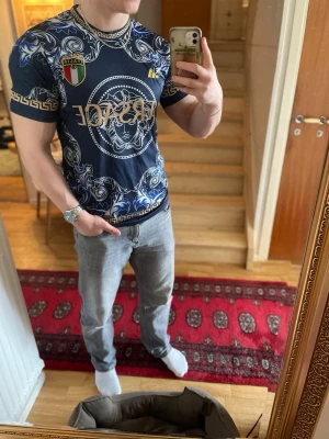 Blå Versace t-shirt med mönster - Säljer en blå t-shirt från Versace med guldiga detaljer och stort Medusa-motiv på bröstet. T-shirten har korta ärmar, tryckta mönster och logotyp framtill. Perfekt för dig som vill sticka ut med en exklusiv look.
