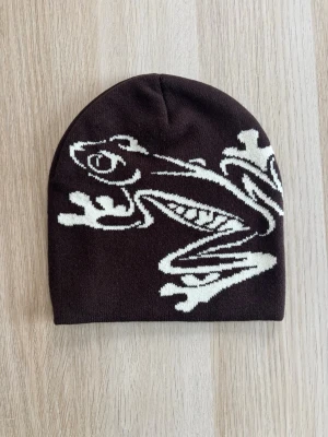 Brun / krämvit Beanie mössa med groda 🐸  - Brun mössa med vitt grodmönster. One size storlek. Ny.  Unisex.  Skickar gärna i bundle.    