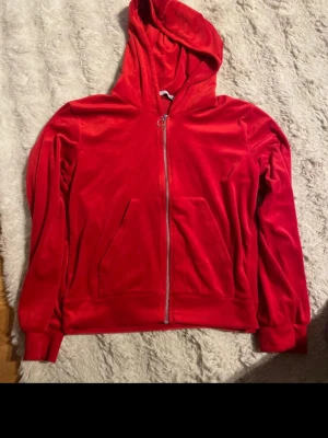 Röd velourhoodie från 157 - Snygg röd hoodie i mjuk velour från 157 med dragkedja framtill och stor känguruficka. Klassisk huva och ribbade muddar vid ärmslut och nederkant. Perfekt för en chill och trendig look.