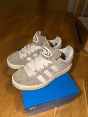 Grå Adidas Campus sneakers i mocka - Snygga Adidas Campus sneakers i grå mocka med vita klassiska tre ränder på sidan och vit sula. I storlek 36. Har ett hål vid hälen 