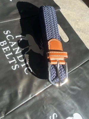 Blått flätat bälte  - Snyggt blått flätat bälte med detaljer i brunt skinn och silverfärgat spänne. Perfekt accessoar för att lyfta din outfit med en stilren touch. Det är 105 cm