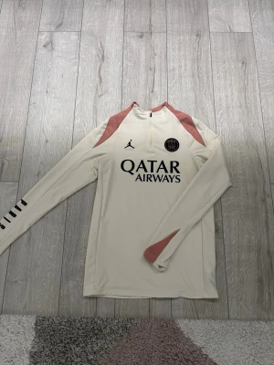 PSG träningsjacka från Jordan - Säljer en snygg långärmad träningsjacka från PSG x Jordan med halv dragkedja. Jackan är ljusbeige med rosa axelpartier. Använd en gång. Pris kan diskuteras! 