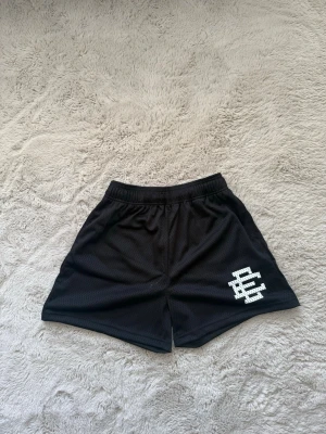 Svarta Eric Emanuel shorts - Säljer ett par ”ÄKTA” svarta Eric Emanuel shorts med vit logga på vänster ben. Shortsen har elastisk midja och är tillverkade i ett luftigt syntetmaterial, perfekta för en sportig och avslappnad stil.