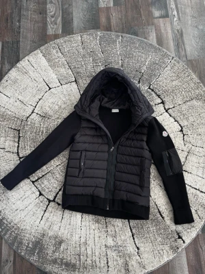 Svart Moncler cardiganjacka med huva - Snygg svart cardiganjacka från Moncler med vadderad front och huva. Ärmarna är ribbstickade och har en patch med Moncler-logga på vänster arm. Två dragkedjefickor framtill och hel dragkedja. Perfekt för lager på lager och streetwear. Priset kan diskuteras vid en effektiv affär. Storleken passar även M
