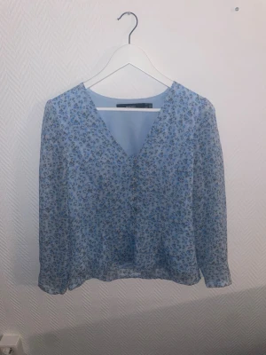 Blommig blå blus från Vero Moda - Söt blus i ljusblått från Vero Moda med småblommigt mönster i blått och brunt. Blusen har v-ringning och långa ärmar i ett tunt, lätt material som känns luftigt och perfekt för vår och sommar. Passar till både jeans och kjol för en fräsch look.