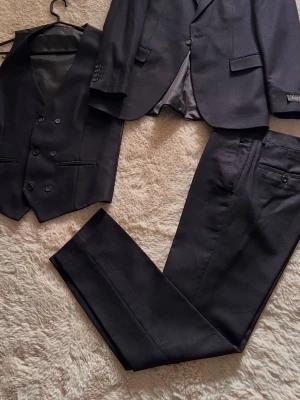 Mörkblå tredelad kostym - Snygg mörkblå tredelad kostym med kavaj, väst och byxor. Kavajen har klassisk passform med knappar vid ärmslut och två fickor. Väst med dubbelknäppning och två fickor framtill. Perfekt för dig som vill ha en stilren och tidlös look.