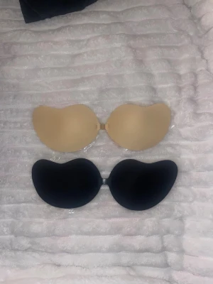 Självhäftande bh i beige och svart - Två självhäftande bhar utan axelband och rygg, en i beige och en i svart. Perfekt för plagg med öppen rygg eller axellöst. Tillverkade i silikonmaterial med klister på insidan för bra fäste. Kopplas ihop framtill med liten spänne.