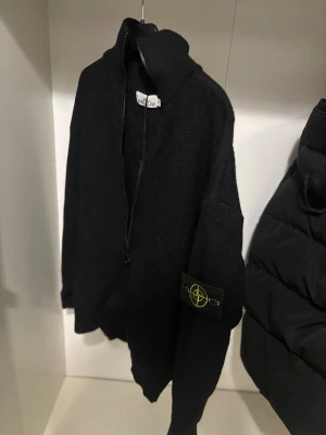 Svart stickad ziptröja Stone Island - Säljer en svart stickad ziptröja från Stone Island med hög krage och dragkedja framtill. Tröjan har den klassiska Stone Island-loggan på ärmen och är tillverkad i ett tjockare, mjukt material. Perfekt för lager på lager och streetwear-stil.