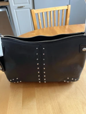 Svart handväska med nitar - Cool svart handväska från Michael Kors i slätt läder med silvriga nitar i raka rader framtill och på hörnen. Väskan har en bred axelrem och dragkedja upptill. Perfekt för dig som gillar en edgy stil och vill ha en väska som sticker ut.